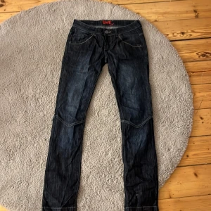 Mörkblå jeans från SWE med detaljer - Mörkblå jeans från SWE med snygga kontrastsömmar och unika bakfickor med knappar och vågiga sömmar. Klassisk femficksmodell och normal passform. Perfekta för dig som vill ha ett par jeans med lite extra attityd.