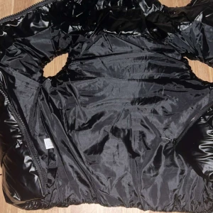 Svart pufferjacka med huva inte använd  - Snygg svart pufferjacka med glansig finish och huva. Jackan har dragkedja framtill och två fickor med dragkedja. Perfekt för kyliga dagar när du vill hålla stilen. Jackan är vadderad och har en modern, lite oversized look.