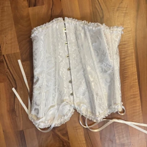 Vit korsett med spets och snörning - Säljer en vit korsett med vackert broderat mönster och glansig finish. Toppen har snörning i ryggen, knäppning fram och volangkant upptill och nedtill. Perfekt för dig som vill ha en statement piece med romantiska detaljer.