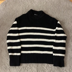 Randig stickad tröja från Gina Tricot - Säljer en svart och vit randig stickad tröja från Gina Tricot i storlek XS. Tröjan har grov ribbstickning, hög hals och långa ärmar. Perfekt för dig som gillar en klassisk men trendig look med lite boxig passform.💗