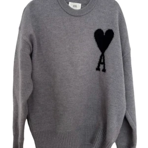 Grå stickad tröja från Ami Paris - Snygg grå stickad tröja från Ami Paris med svart hjärtmönster och bokstaven A på bröstet. Tröjan har rund hals, ribbade muddar och lång ärm. Perfekt för dig som gillar stilrena detaljer och vill ha en chill look.