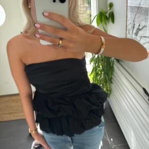 Svart volangig tubtopp - Säljer en svart axelbandslös tubtopp med volanger och rynkade detaljer. Toppen har en bandeauformad överdel och är tillverkad i ett lätt tyg som ger en luftig känsla. Perfekt att styla med jeans för en trendig look.