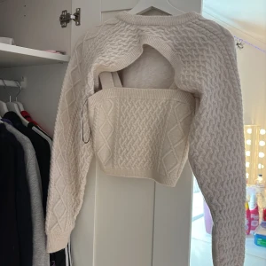 Beige croppad stickad tröja - Säljer en beige croppad stickad tröja med snyggt kabelmönster och öppen rygg. Tröjan har långa ärmar och ribbade muddar. Perfekt för dig som vill ha en trendig och unik look med coola detaljer. (Sett med 2 delar) 