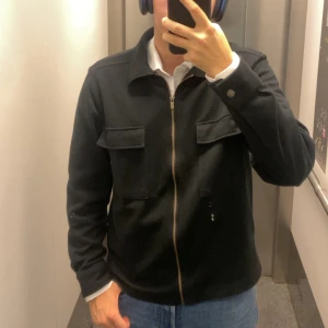 Svart overshirt - Svart overshirt med stilren design och två stora fickor framtill. Jackan har en rak passform, klassisk krage och stängs med en metallfärgad dragkedja. Materialet känns mjukt och har en lätt struktur, perfekt för lager-på-lager. Långärmad och enkel att matcha med jeans.