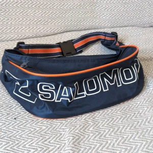 Vintage Blå mag/axelväska från Salomon - Snygg blå axelväska som också går att ha som magväska från Salomon med stort vitt logotryck och orange detaljer. Väskan har justerbart band med ränder i blått och orange samt dragkedja upptill. Perfekt för att bära med sig det viktigaste på språng. 26x11x10cm 🌷ALLT MÅSTE BORT INNAN 5 OKTOBER BUDA GÄRNA 🌷