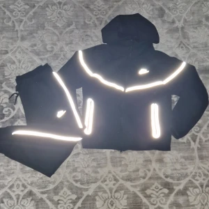 Nike tech tracksuit reflective - Svart hoodie från Nike med dragkedja och huva, prydd med feta reflexdetaljer på bröstet och vid fickorna. Tillverkad i mjuk bomullsmix, perfekt för dig som vill sticka ut. Matchande byxor med reflexränder och Nike-logga ingår.