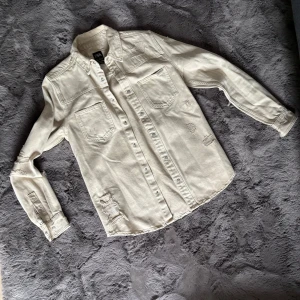 Beige jeansskjorta från ZARA - Säljer en beige jeansskjorta från ZARA med slitna detaljer och två bröstfickor. Skjortan har knappar hela vägen och lång ärm med knapp vid mudden. Perfekt för dig som gillar en avslappnad och cool stil.