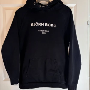 Svart hoodie från Björn Borg - Svart hoodie från Björn Borg med vit text på bröstet. Klassisk känguruficka framtill, huva med snörning och ribbade muddar. Perfekt för chill dagar eller när du vill ha en enkel och snygg look.