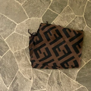 Fendi brun och svart halsduk - Snygg och lång halsduk från Fendi med ikoniskt FF-mönster i svart mot brun bakgrund. Halsduken har fransar i ändarna och är perfekt att svepa runt halsen för en lyxig touch. Materialet känns mjukt och skönt mot huden.