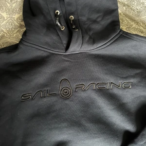 Marinblå Sail Racing hoodie med brodyr - Marinblå hoodie från Sail Racing med broderad logga på bröstet. Tröjan har huva med snörning och en stor magficka. Mjuk insida och ribbade muddar vid ärmslut och nederkant. Perfekt för dig som gillar stilren streetwear. Använd en gång. Passar både tjej och kille