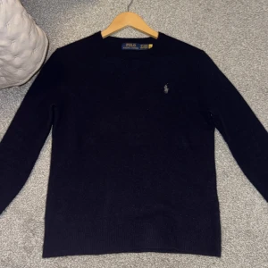 Mörkblå stickad tröja Polo Ralph Lauren - Mörkblå stickad tröja från Polo Ralph Lauren med klassisk rund hals och broderad logga på bröstet. Tröjan har lång ärm och är i en mjuk, skön ullblandning. Perfekt för dig som gillar stilren och tidlös design.