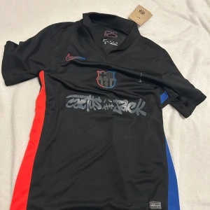 FC Barcelona x Nike Lamine Yamal tröja - Snygg svart FC Barcelona fotbollströja från Nike med Lamine Yamal och nummer 10 på ryggen. Tröjan har röda och blå detaljer på sidorna, klubbmärke på bröstet och 'Cactus Jack' tryck framtill. Tillverkad i lätt och ventilerande material.