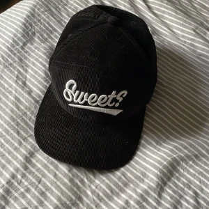 Svart manchesterkeps från Sweet - Svart keps i manchester med vit broderad Sweet?-logga framtill. Kepsen har klassisk snapback-spänne bak och grön undersida på skärmen. Perfekt accessoar för en cool streetstil.