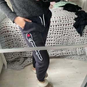 Mörkblå mjukisbyxor från Champion - Mörkblå mjukisbyxor från Champion med vit snörning i midjan och stor broderad logga längs ena benet. Byxorna har mudd i benslutet. Barnstorlek 2X-Large (174/179 cm) men passar XS/S. 