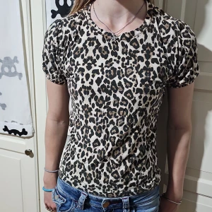 Leopardmönstrad topp Young Wear - Snygg kortärmad topp från Young Wear med leopardmönster i beige, svart och brunt. Modellen har puffiga ärmar med resår och rund halsringning. Perfekt för dig som vill sticka ut med ett djurmönstrat plagg. Mjuk och skön känsla, passar till jeans eller kjol. Lite tvättpåverkad