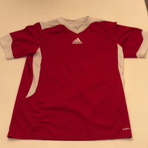 Röd Adidas fotbollströja Climalite - Röd fotbollströja från Adidas med vita detaljer på axlar och sidor, samt Adidas-logga på bröstet. Tillverkad i lätt och ventilerande Climalite-material som passar perfekt för träning eller match. Klassisk V-ringning och korta ärmar.