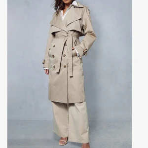 Beige trenchcoat från Misspap - Snygg beige trenchcoat från Misspap med klassisk krage, knappar framtill och bälte i midjan. Jackan har långa ärmar med spänne vid mudden och en omlott-design som ger en cool siluett. Perfekt för dig som gillar stilrena och tidlösa plagg.