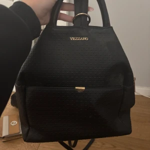 Svart ryggsäck från Vezzano - Snygg svart ryggsäck från Vezzano med gulddetaljer och strukturerad yta. Praktiskt ytterfack framtill och stilrena handtag upptill. Perfekt för dig som vill ha en stilren och funktionell väska till vardagen.