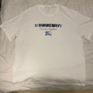 Vit Burberry t-shirt med svart/blått tryck - Vit t-shirt från Burberry med blått tryck på bröstet där det står 'Burberry London, England' och en klassisk riddarlogga. T-shirten har rund hals och korta ärmar, perfekt för en clean och stilren look.