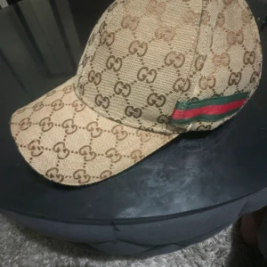 Gucci keps med GG-mönster och röd/grön detalj - Snygg beige keps från Gucci med klassiskt GG-monogram över hela ytan och ikonisk röd/grön rand på sidan. Justerbar rem baktill i brunt skinn.