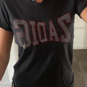 Svart t-shirt med ZADIG-tryck - Svart t-shirt från Zadig & Voltaire med glittrande ZADIG-tryck i stenar framtill. Klassisk rund halsringning och korta ärmar. Perfekt för dig som gillar statement-plagg med lite edge.