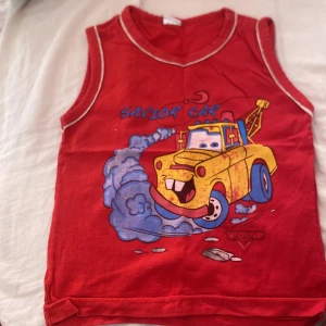 Röd Cars linnetopp med tryck - Säljer en röd ärmlös linnetopp med motiv från Cars och texten 'Savior Car' framtill. Toppen har kontrasterande vita sömmar runt hals och ärmhål samt en stor färgglad bild på en gul bärgningsbil. Perfekt för dig som gillar animerade filmer och coola tryck.