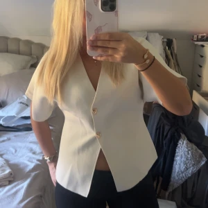 Vit blus med knappar från Zara - Säljer en stilren vit blus från Zara med korta ärmar och v-ringning. Blusen har två snygga knappar framtill och är lätt figursydd för en clean look. Materialet är mjukt och har en lätt struktur. Perfekt för dig som gillar minimalistisk stil. Knappt använd