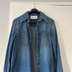 Jeansskjorta från Calvin Klein Jeans - Säljer en klassisk blå jeansskjorta från Calvin Klein Jeans med två bröstfickor och snygga kontrastsömmar. Skjortan har tryckknappar framtill och en avslappnad passform. Storlek L, använd 1 gång. 