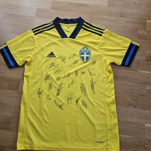 Svenska herrlandslagets signerade matchtröja - Gul fotbollströja från Adidas med blå detaljer, officiellt Svenska landslagets emblem på bröstet och flera autografer på framsidan. Tröjan är tillverkad i lätt och ventilerande Aeroready-material och har korta ärmar samt klassiska Adidas-ränder på axlarna.