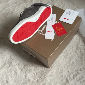 Grå mockasneakers från Louboutin - Säljer ett par grå sneakers från Christian Louboutin i mjuk mocka med klassisk röd sula. Skorna har rund tå, snörning och vita gummisulor. Kommer med originalkartong och äkthetskort. Perfekta för dig som vill ha en lyxig och stilren look.