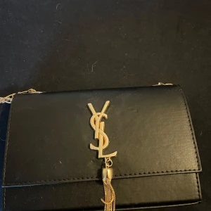 Svart YSL axelväska med guldkedja - Snygg svart axelväska från YSL i slätt skinn med guldfärgad kedja och ikonisk YSL-logga framtill. Väskan har ett elegant hängande gulddetalj och stilren design som ger en lyxig känsla. Perfekt storlek för det viktigaste och enkel att bära över axeln.  Pris går att diskuteras. 