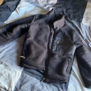 Svart teddyjacka från Canada Goose - Säljer en svart teddyjacka från Canada Goose med patch på ärmen. Jackan har dragkedjor på fickorna, hög krage och detaljer i nylon på bröstet. Perfekt för kalla dagar och har en riktigt skön och mjuk känsla.