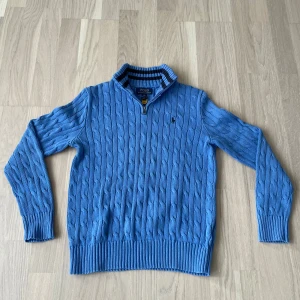 Blå kabelstickad tröja Polo Ralph Lauren - stickad blå tröja från Polo Ralph Lauren med halv dragkedja och ribbade muddar. Tröjan har svart broderad logga på bröstet och krage med svarta ränder på insidan. Tillverkad i mjuk bomull och perfekt för lager-på-lager.
