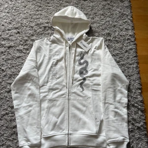 Vit hoodie med ormmotiv från Trapper - Vit hoodie från Trapper med dragkedja, huva och snörning. Framsidan har ett coolt ormmotiv i svarta stenar och Trapper-logga i glittriga stenar. Mjuk bomullskänsla och känguruficka framtill. Perfekt för dig som gillar streetstyle.