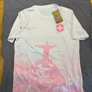 Rosa Brasilien fotbollströja  - Snygg vit fotbollströja från Nike med rosa detaljer och tryck av Kristusstatyn i Rio de Janeiro. Bröstet har CBF-logga och Nike swoosh i rosa. Tröjan är kortärmad och tillverkad i ett lätt, ventilerande material som passar perfekt för träning eller match.