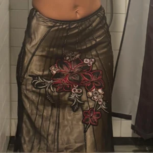 Långkjol med broderade blommor - Snygg långkjol i mörkbrunt transparent tyg med stora broderade blommor i rött, svart och rosa framtill. Kjolen har en elegant och unik look med tunna svarta linjer som detaljer. Perfekt för dig som vill sticka ut med en cool och kreativ stil.
