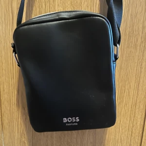 Svart axelväska från BOSS Parfums - Stilren svart axelväska från BOSS Parfums i slätt skinnliknande material. Väskan har justerbar axelrem, silverfärgad dragkedja och logga framtill. Perfekt för att bära det viktigaste med en clean look.