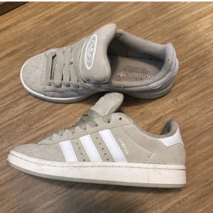 Adidas Campus grå sneakers mocka - Grå Adidas Campus sneakers i mocka med vita klassiska ränder på sidorna och vit logga på plösen. Skorna har rund tå, platt sula och snörning. Perfekta för dig som gillar en clean och stilren look med retrovibbar.