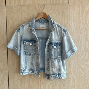 Ljusblå croppad jeansjacka s.Oliver - Snygg ljusblå croppad jeansjacka från s.Oliver med korta ärmar och klassisk krage. Jackan har två bröstfickor med knappar och uppvikta ärmslut. Perfekt för dig som vill ha en chill och trendig look.