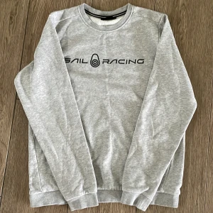 Grå Sail Racing sweatshirt - Snygg grå sweatshirt från Sail Racing med svart logga framtill. Tröjan har rund halsringning, långa ärmar och ribbade muddar vid ärmslut och nederkant. Perfekt för dig som gillar stilren och sportig look.