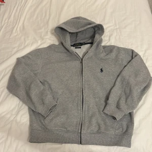 Grå hoodie från Polo Ralph Lauren - Grå hoodie från Polo Ralph Lauren med dragkedja och klassisk blå broderad logga på bröstet. Tröjan har huva, ribbade muddar och fickor framtill. Perfekt för chill dagar och enkel att matcha med jeans eller joggers. Har krympt till Xs