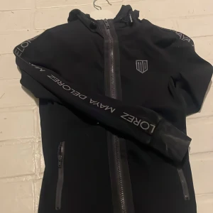 Svart vindjacka från Maya Delorez - Svart vindjacka från Maya Delorez med huva och hel dragkedja framtill. Jackan har grå detalj längs ryggen och logga på bröstet. Snygga tryck på ärmarna och två fickor med dragkedja. Perfekt för dig som gillar sportig stil. Använd fåtal gånger, fint skick. Nypris: 1000kr 