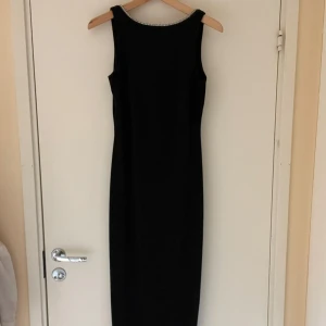 Svart långklänning med stendetalj - Elegant svart långklänning med ärmlös design och rund halsringning och djup rygg. Den är dekorerad med glittrande stenar och har en slits i sidan.