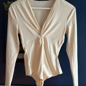 Beige body med v-ringning - Snygg beige omlott body med lång ärm och djup v-ringning framtill. Twistad detalj vid bysten och är tillverkad i ett mjukt, stretchigt material som sitter skönt på kroppen. Perfekt för dig som gillar stilrena och trendiga plagg.