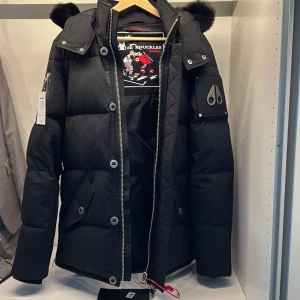 Svart pufferjacka från Moose Knuckles - Säljer en svart pufferjacka från Moose Knuckles med fet huva och pälskant. Jackan har stora fickor med knappar, dragkedja och tryckknappar framtill samt logga på ärmen. Perfekt för kalla dagar och riktigt snygg streetstil. Helt ny har aldrig använt den har alla tags kvar och kvitton. Köpt från NK i Stockholm