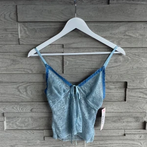 Ljusblå spetslinne från Nelly - Supersöt ljusblå linne-topp från Nelly i transparent spets med tunna axelband och dekorativ knytning framtill. Toppen är oanvänd.