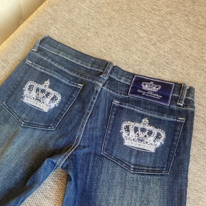 Victoria Beckham jeans med krona Bootcut Lågmidjade - Mörkblåa Victoria Beckham jeans💙De är lågmidjade och bootcut. Små diamanter på bakfickorna💎Storlek: S💘 Väldigt fint skick och har endast använts ett fåtal gånger. Passar perfekt för dig som gillar unika vintage jeans! De matchar med allt😍