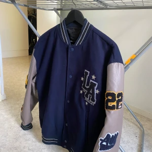 Blå och grå college jacka - Snygg bomberjacka i college-stil från ASOS DESIGN. Jackan är marinblå med gråa ärmar i skinnimitation, broderade LA- och California-detaljer samt gula och vita applikationer. Ribbstickad krage och muddar med ränder, knäppning framtill. Storlek S men ganska stor i storlek