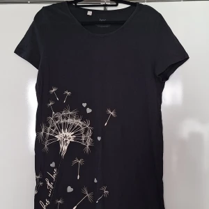 Svart kortklänning/lång t-shirt,  med maskrosmotiv L - Svart kortklänning från bpc med rund hals och korta ärmar. Klänningen har ett snyggt maskrosmotiv och små hjärtan i vitt och grått på framsidan. Tillverkad i mjuk bomull, perfekt för en chill och avslappnad stil.