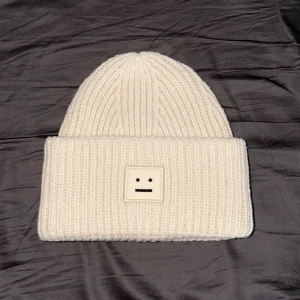 Vit stickad mössa från Acne Studios - Vit ribbstickad mössa från Acne Studios med bred uppvikt kant och det ikoniska ansiktsloggan framtill. Tillverkad i mjuk ull som håller dig varm och snygg under kalla dagar. Perfekt accessoar för en clean och stilren look.